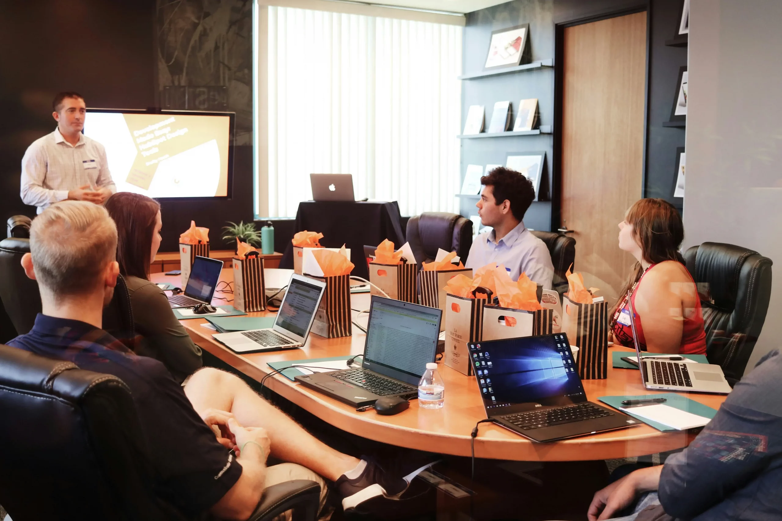 A professional IQAC team meeting where a speaker stands at the head of a conference table presenting to a group of seated colleagues. Everyone has open laptops, and the modern office room features large windows showing a city view.